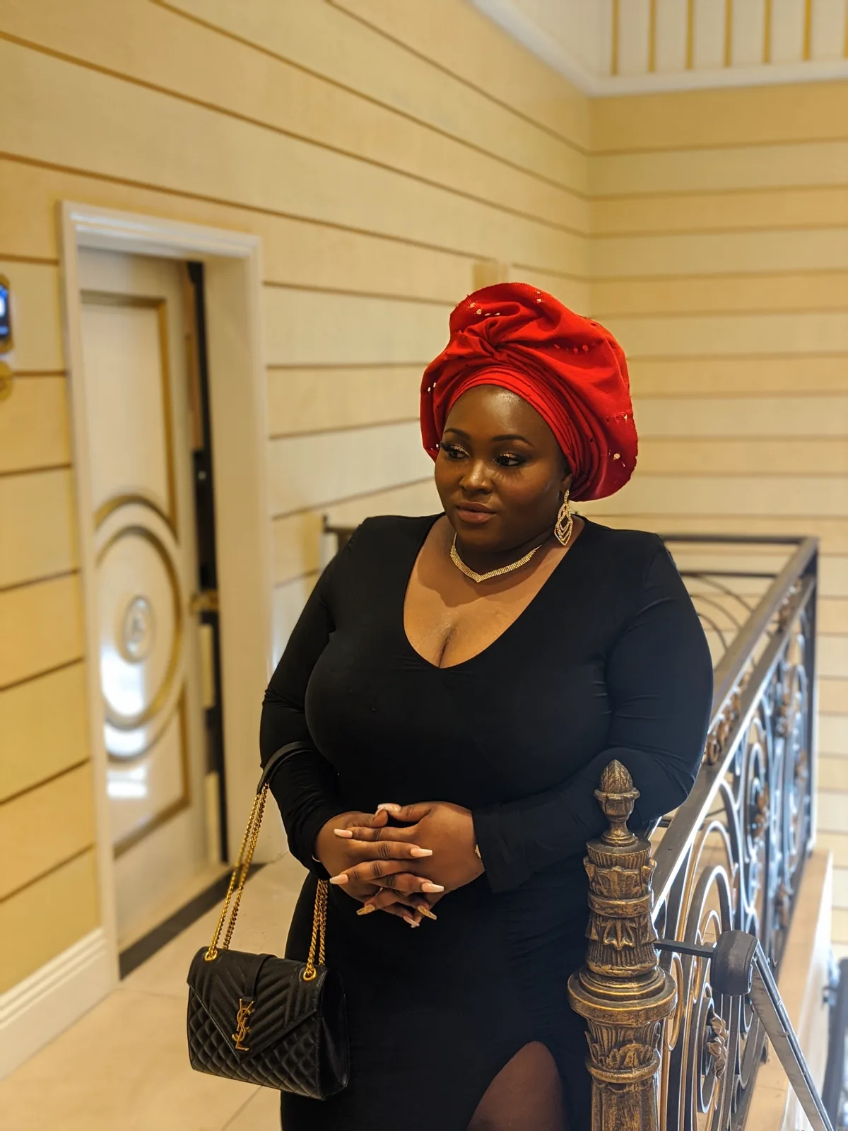 Chizo in a red head wrap and black dress, standing beside a staircase railing.