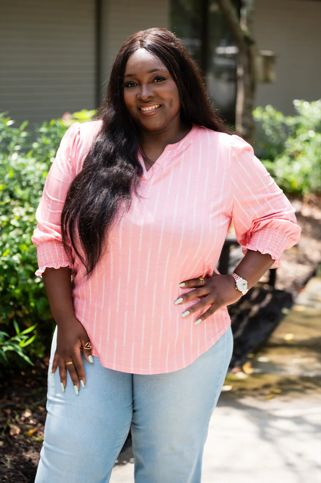 Chizo in a pink blouse and light jeans, standing in a garden with one hand on her hip.